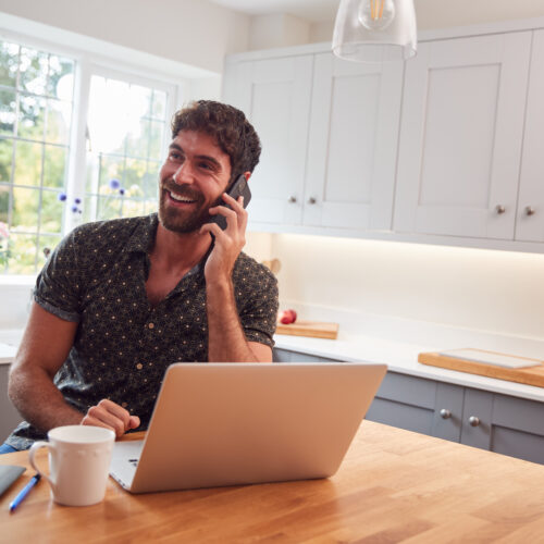 Man In Kitchen With Mobile Phone Working From Home Using Laptop During Health Pandemic