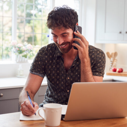 Man In Kitchen With Mobile Phone Working From Home Using Laptop During Health Pandemic